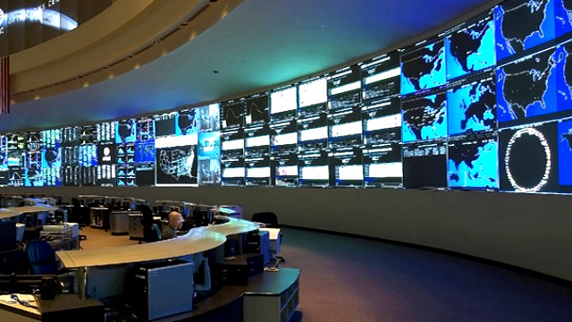 A wall full of monitors, with people monitoring from a desk in the room