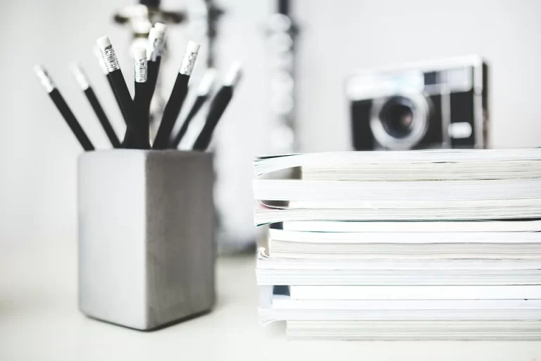 A black and white photo of a pencil holder full of pencils, and a stack of books with a camera on top of them.